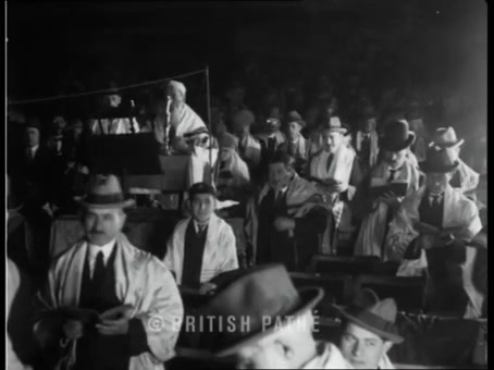 British Pathé frame: crowded hall of men in hats and talleisim, bimah in the background