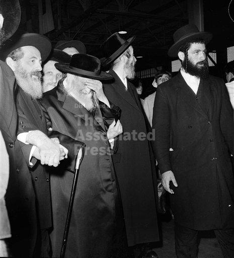 The Satmar Rebbe, Reb Yoel Teitelbaum, at the Queen Mary pier on August 31, 1965, hand over his face, with men in black hats under the terminal roof