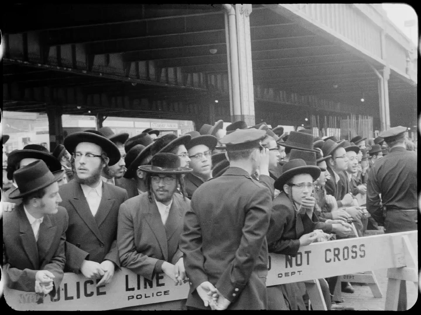 Satmar chassidim in black hats stand behind a police line under the pier shed