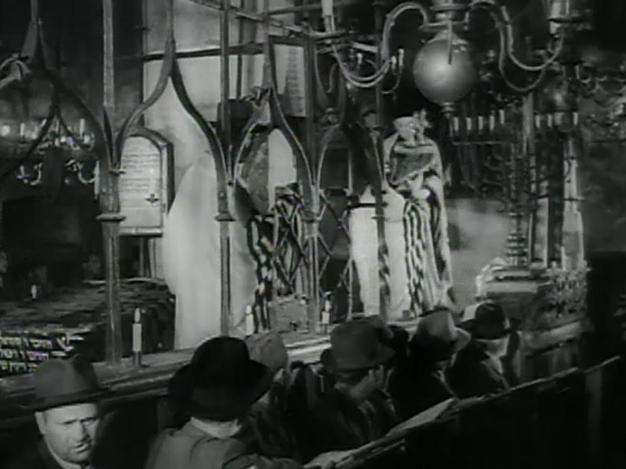 1946 film frame: Torah ceremony inside the Old-New Synagogue, wrought-iron bimah cage, men in hats and talleisim